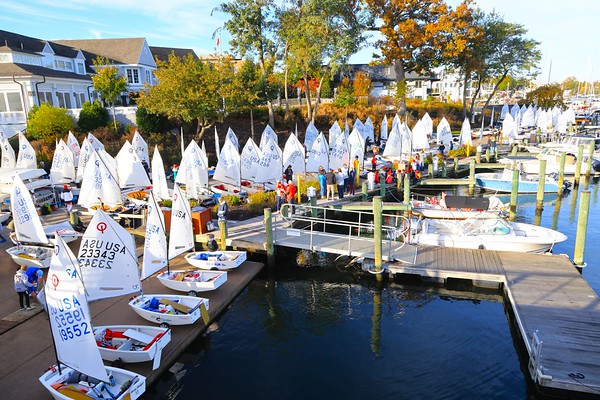 Sailboats with white sails are lined up at a marina dock. Many people, some in red jackets, stand nearby. Trees with autumn leaves and houses are visible in the background under a bright sky.