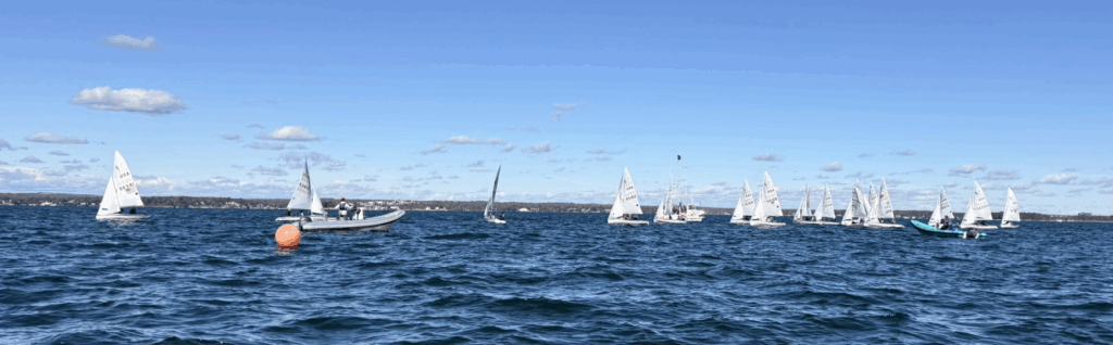 A group of small sailboats races on a blue lake under a clear sky, with a few motorboats nearby and a bright orange buoy floating in the water. A distant shoreline is visible in the background.