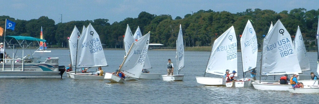 A group of small sailboats with children sailing near a docked pontoon boat on a lake. Trees line the shore in the background under a partly cloudy sky.