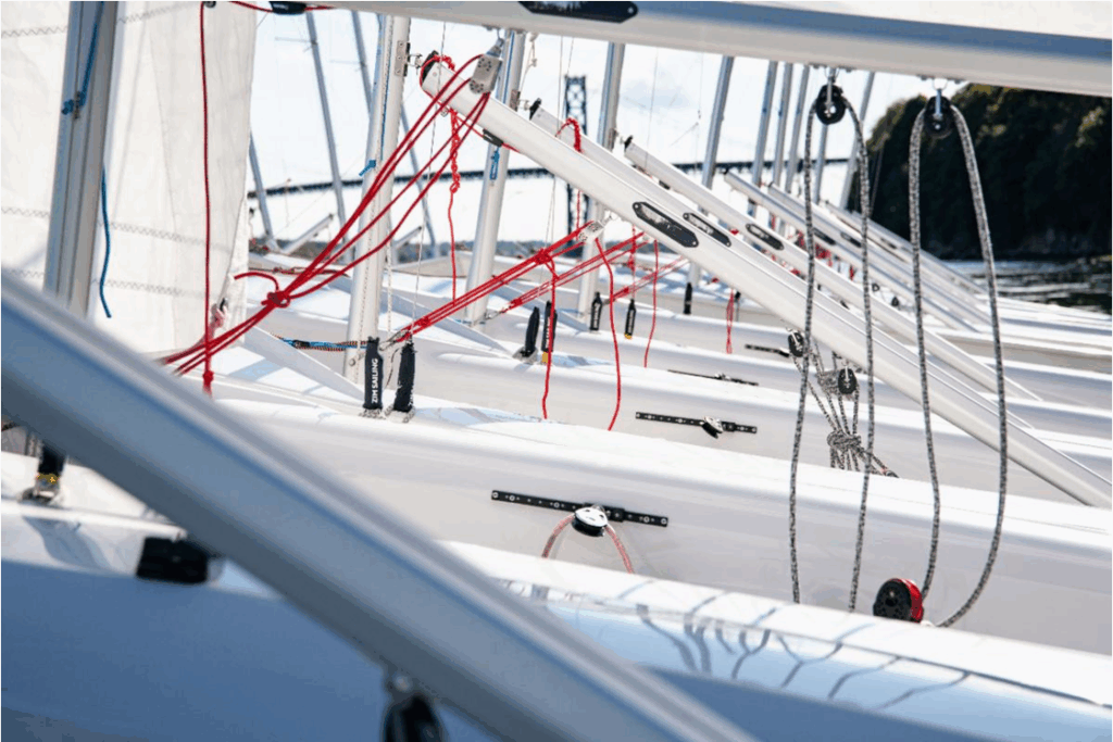 A close-up view of several white sailboats lined up side by side, showing their angled masts, ropes, and rigging, with a blurred natural background and water visible.