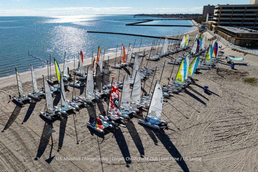 Aerial view of numerous multihull sailboats with colorful sails lined up on a sandy beach by calm water, next to buildings, at the U.S. Multihull Championship in Corpus Christi, Texas.