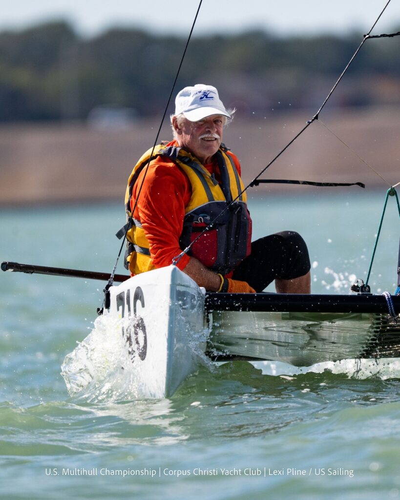 An older man in an orange shirt, yellow life jacket, and white cap sails a small catamaran on the water during a multihull sailing championship. The sailboat splashes through the waves under a sunny sky.