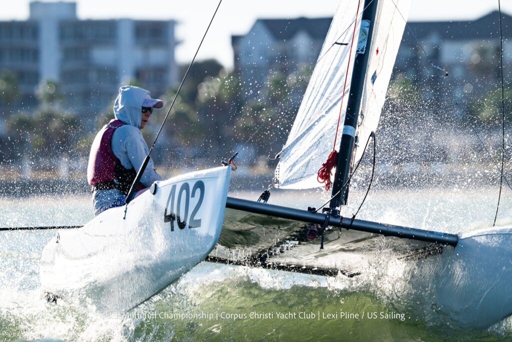 A sailor wearing a hoodie and sunglasses navigates sailboat number 402 through splashing water during a race, with blurred buildings and palm trees in the background.