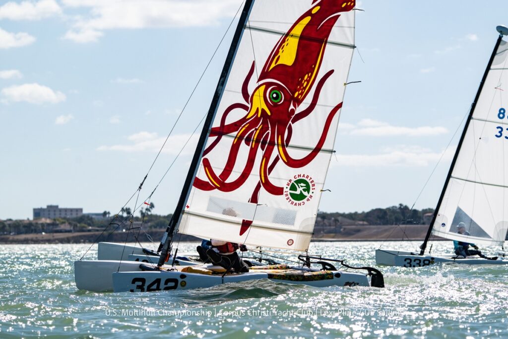 A sailboat with a large, colorful squid design on its sail glides through the water during a race, with another sailboat in the background and a shoreline visible under a bright, sunny sky.