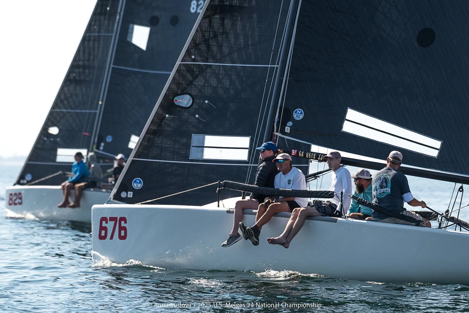 Sailboats competing in a race on calm water, with crew members sitting on the edge of the boats. The sails are black, and boat number 676 is at the forefront, followed closely by other boats.