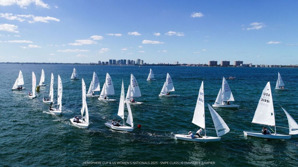 A fleet of small sailboats with white sails races across a blue body of water under a sunny sky, with a city skyline visible in the distance.