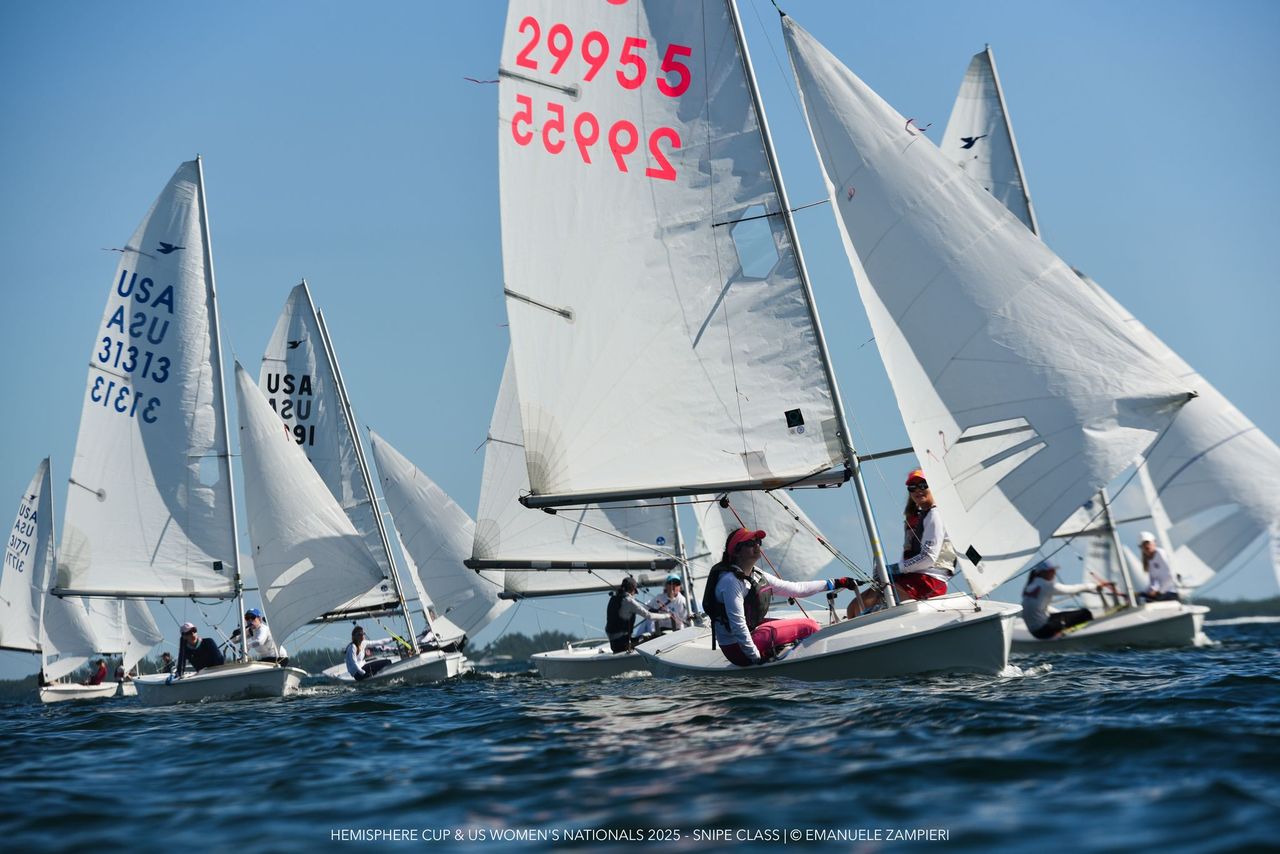 A group of women sailing small sailboats with white sails marked with numbers and “USA” on a sunny day during a race on calm waters. Blue sky and distant trees are visible in the background.