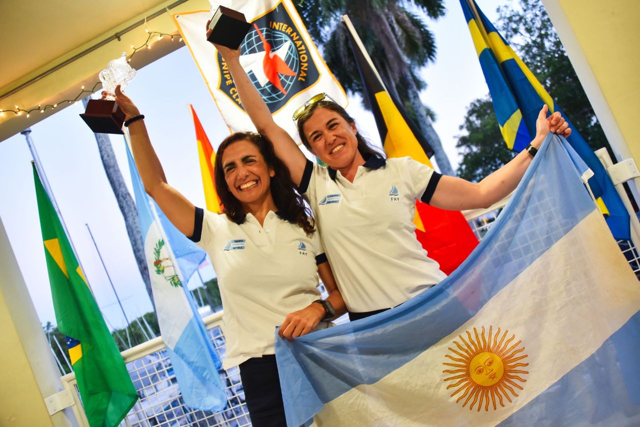 Two women in white polo shirts smile and celebrate with trophies, holding an Argentine flag. Several international flags are in the background, suggesting a competition or awards event.