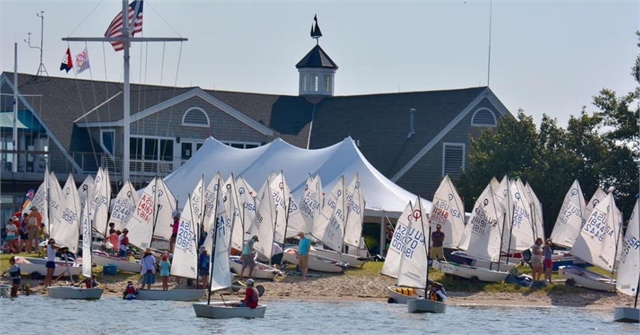 A group of small sailboats with white sails gathers near a sandy shore in front of a building with an American flag. People, including children, prepare boats and socialize under a large white tent.