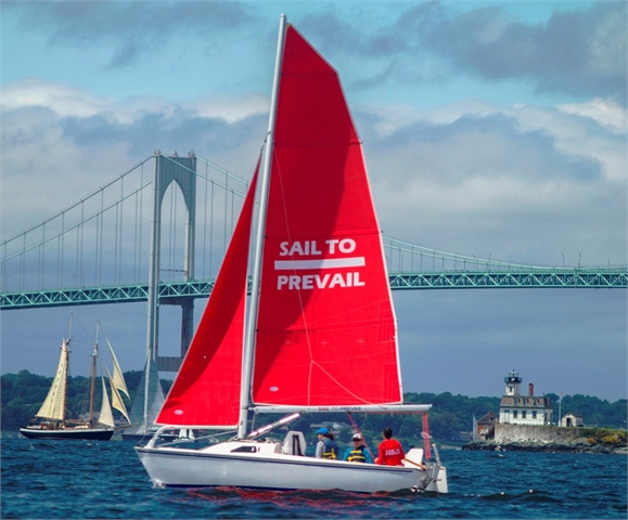 A white sailboat with bright red sails reading 'SAIL TO PREVAIL' sails on the water. Three people are onboard. In the background are historic ships, a bridge, a small lighthouse, and a partly cloudy sky.