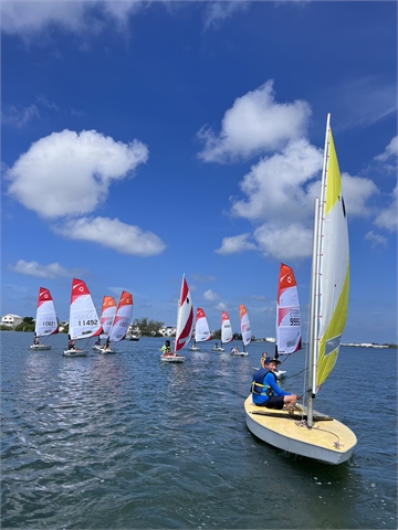 A group of small sailboats with colorful sails race on a sunny lake under a blue sky with scattered clouds. Two people sit in a yellow boat in the foreground. White buildings and trees are visible in the background.