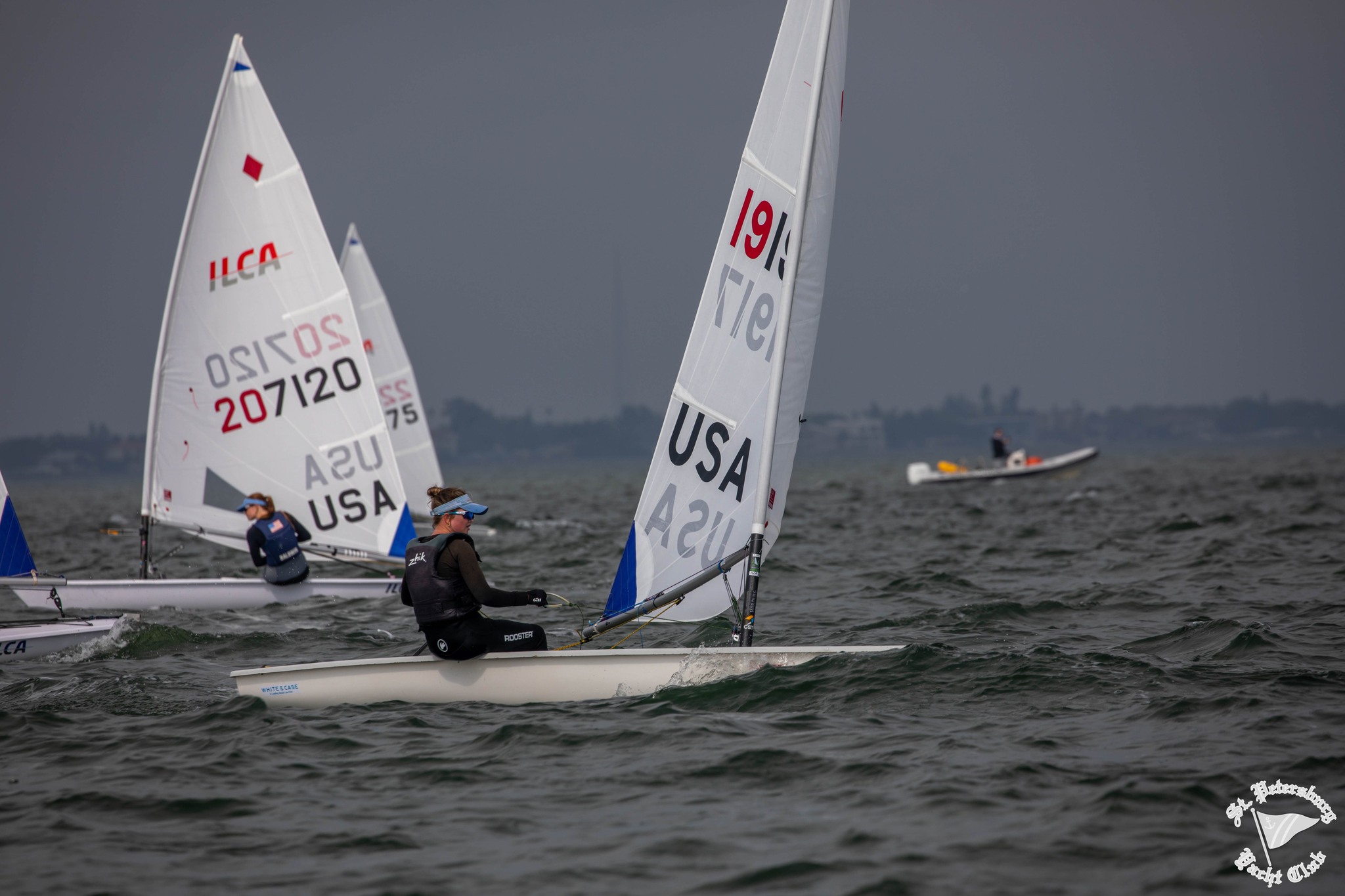Three sailboats with "USA" sails race on choppy water under a gray sky. Sailors wear life jackets and headbands. A distant motorboat and shoreline are visible in the background. St. Petersburg Yacht Club logo is at the bottom right.