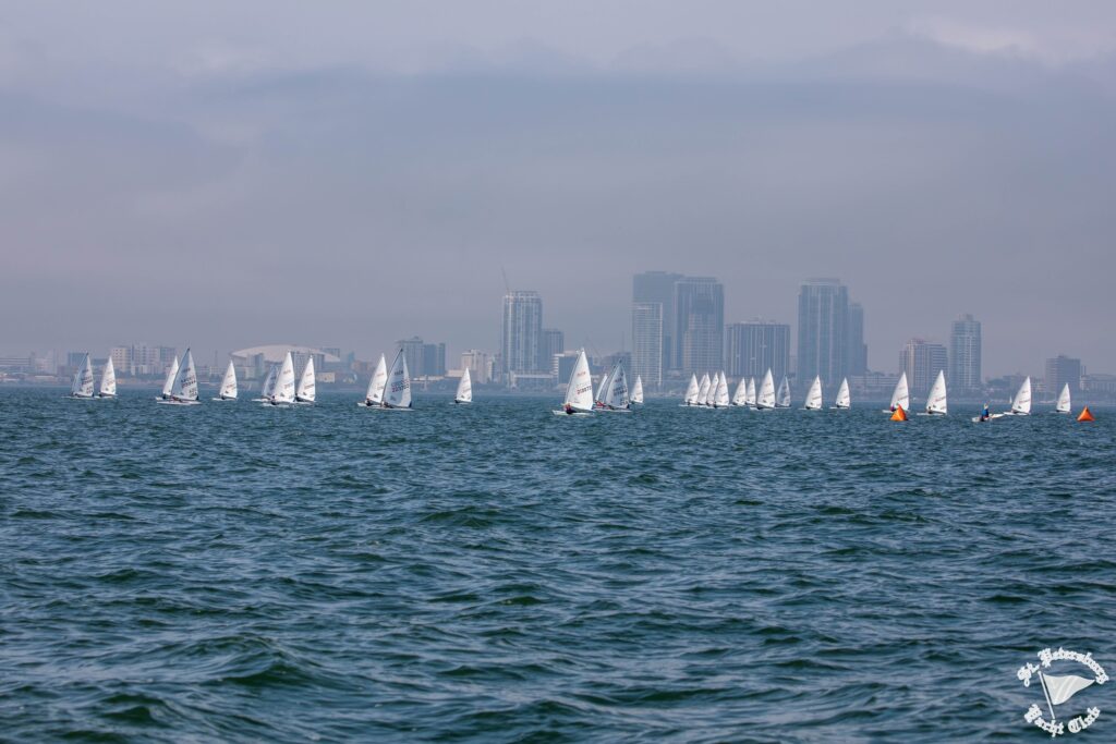 Sailboats race on choppy water under a cloudy sky, with a city skyline and tall buildings visible in the distance. Orange buoys float in the water, and a logo is in the bottom right corner.
