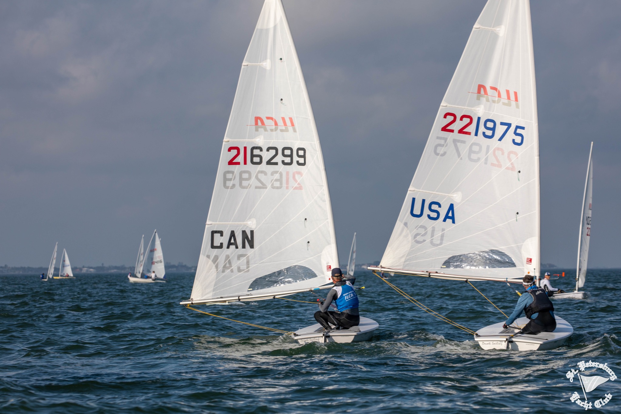 Two sailors race Laser sailboats on open water, with "CAN 216299" and "USA 221975" visible on the sails. Other sailboats are seen in the distance under a partly cloudy sky.