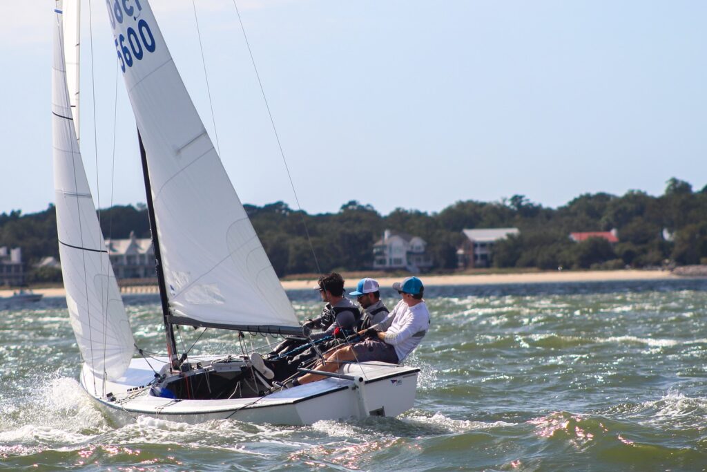 Three people wearing blue caps are sailing a small white sailboat on choppy water near a shoreline with trees and houses visible in the background under a clear sky.