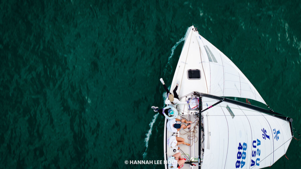 Aerial view of a white sailboat with several people on board sailing on deep green water. The boat's sail has “USA” and “488” written on it.
