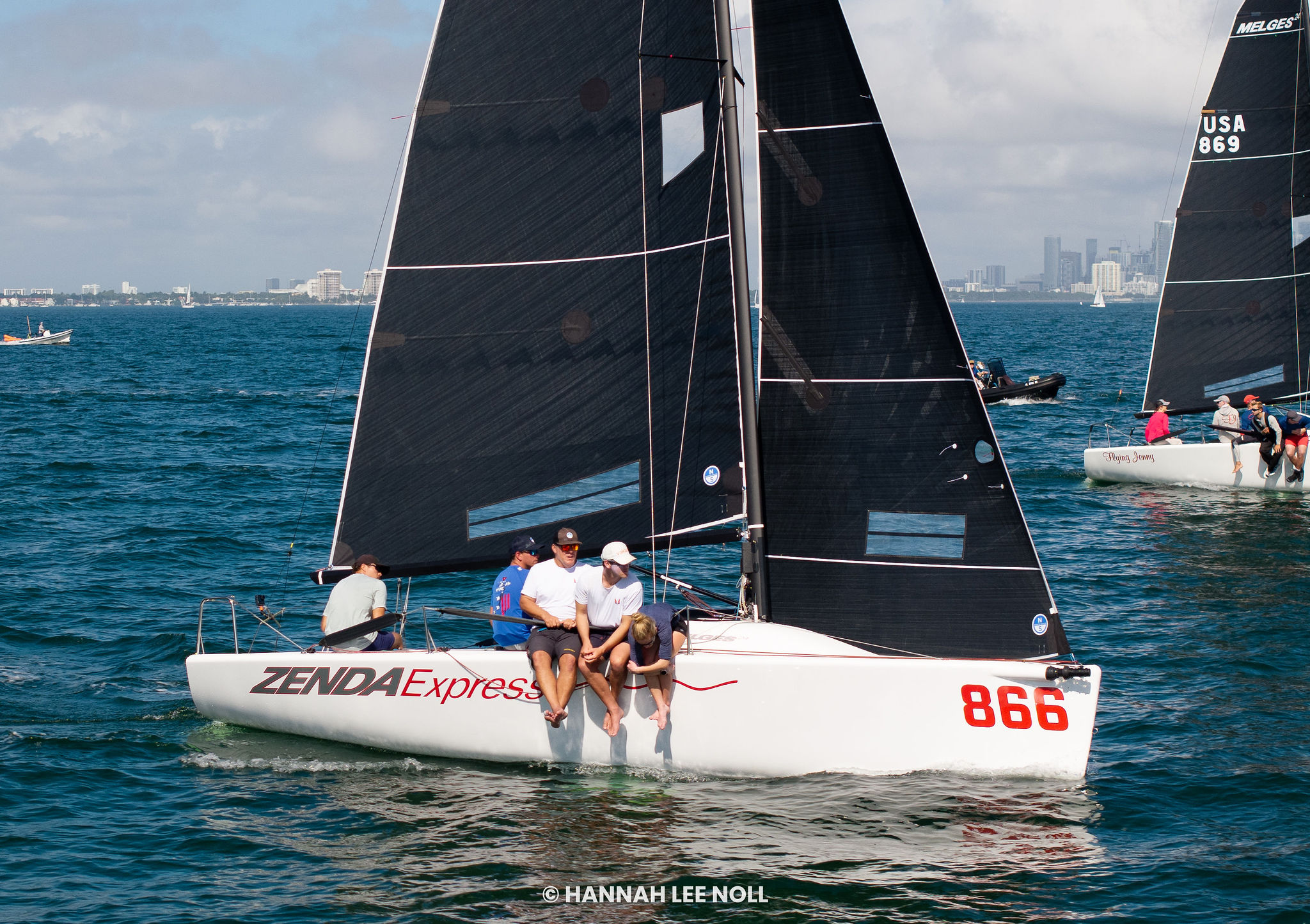 Four people sit on the edge of a white sailboat named "Zenda Express" with the number 866, sailing in blue water with other boats nearby and a city skyline in the background.