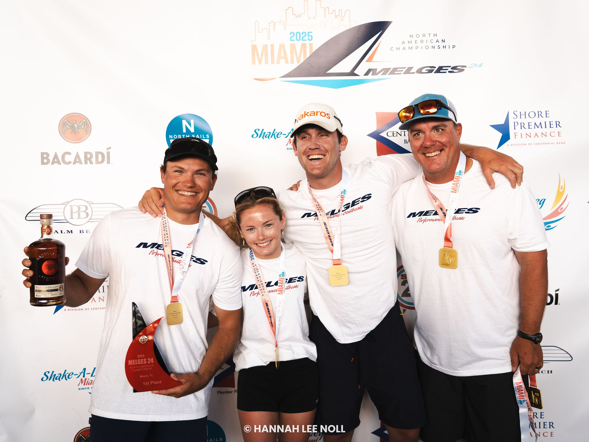 Four smiling people in matching white shirts and medals pose together at a sailing event, holding trophies and a bottle, in front of a branded event backdrop.