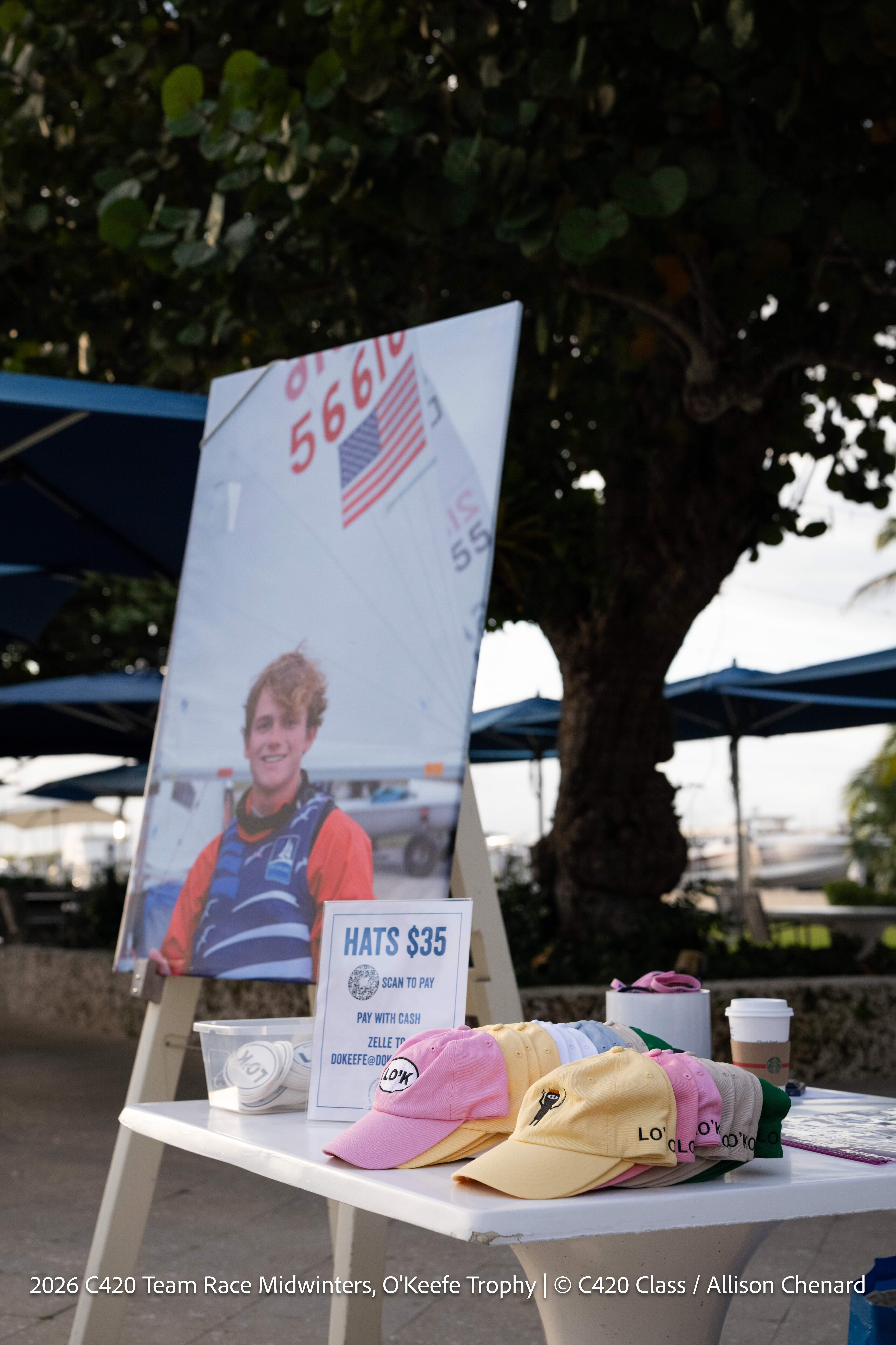 A display table with pastel hats for sale and a sign reading "Hats $35" stands in front of a large photo of a smiling sailor, with trees and sailboats in the background.