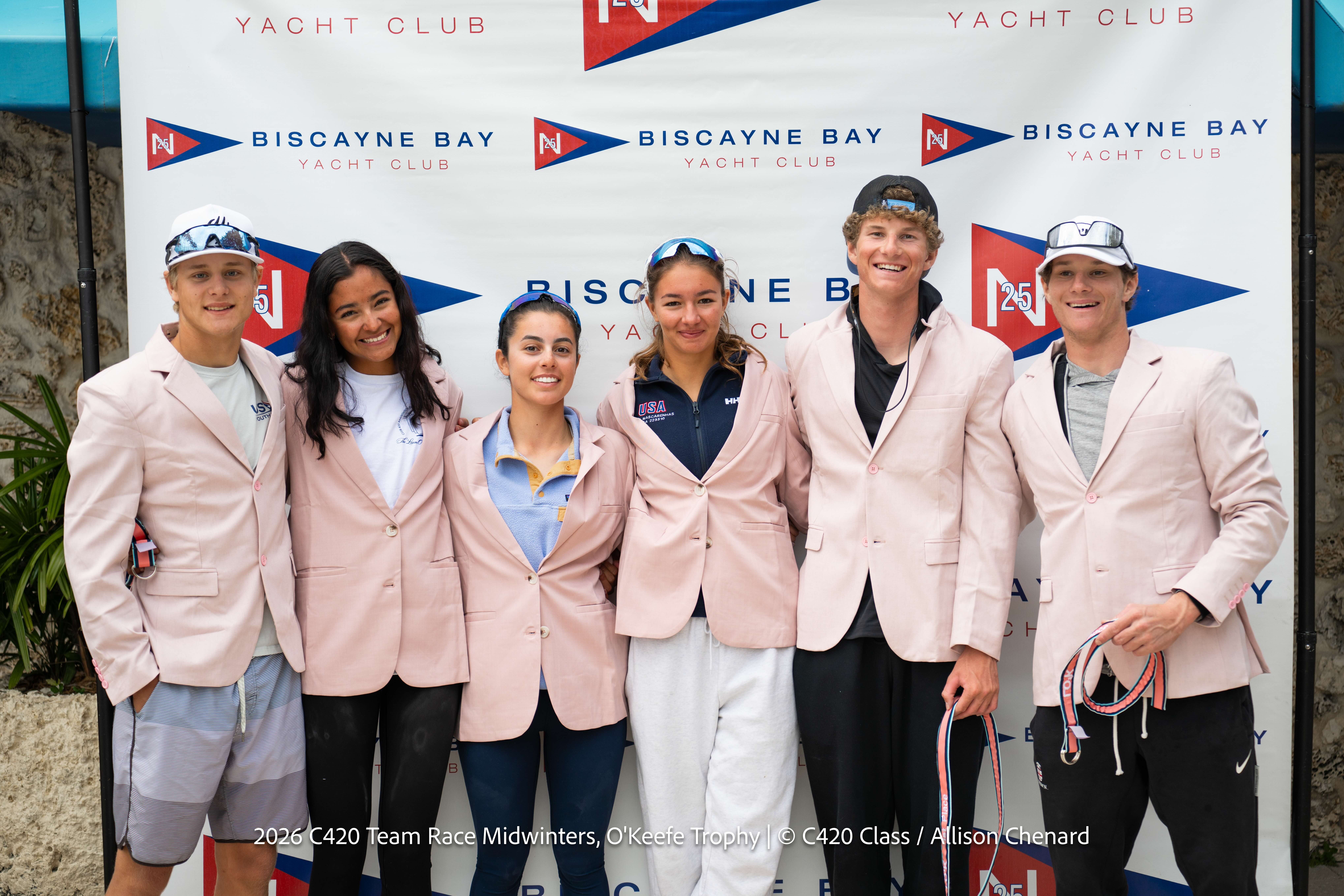 Six young sailors wearing light pink blazers stand smiling in front of a Biscayne Bay Yacht Club banner, posing together with their arms around each other. Some are wearing hats or sunglasses.