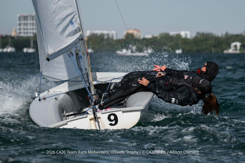 Two sailors in dark gear lean out of a small white sailboat marked with a '9' as water splashes around them during a race on a sunny day, with buildings and trees visible in the background.