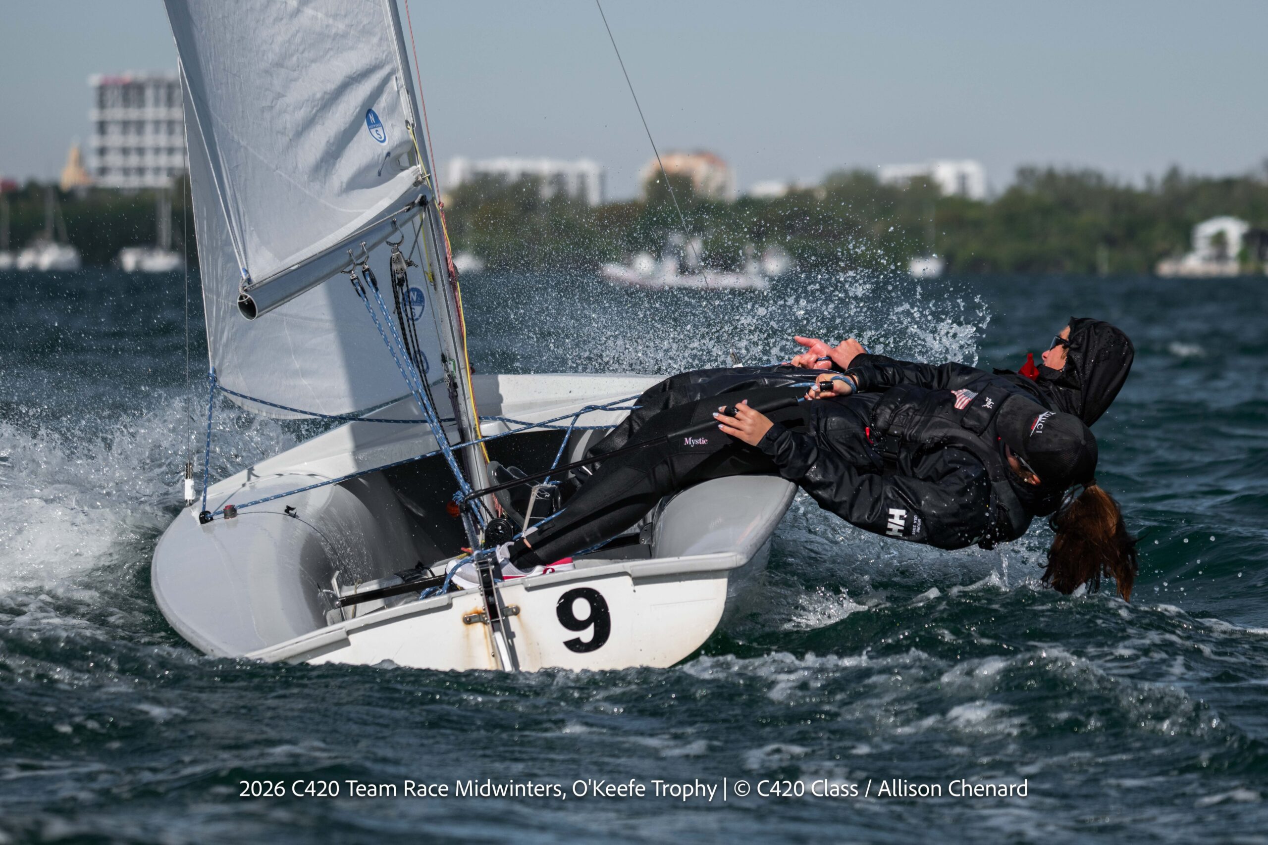 Two sailors in dark gear lean out of a small white sailboat marked with a "9" as water splashes around them during a race on a sunny day, with buildings and trees visible in the background.
