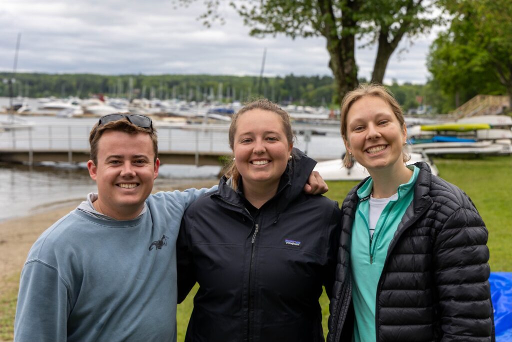 Three smiling people stand close together outdoors near a lake with boats and docks in the background. They are dressed in casual jackets and appear happy, enjoying the day by the waterfront. Trees and grass are visible around them.