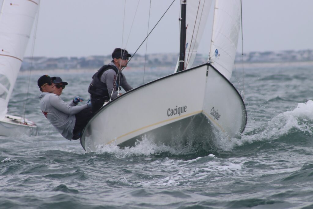 Two people are sailing a boat named "Cacique" through choppy waters, leaning out over the side to balance the vessel. The coastline and another sailboat are visible in the background.