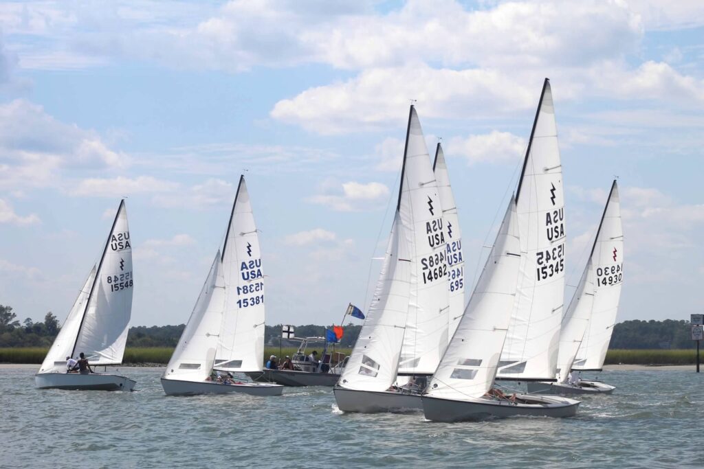 Several small sailboats with white sails, marked 'USA,' compete in a race on a sunny day. The boats gather near a buoy and a small motorboat with flags, with calm water and grassy shoreline in the background.