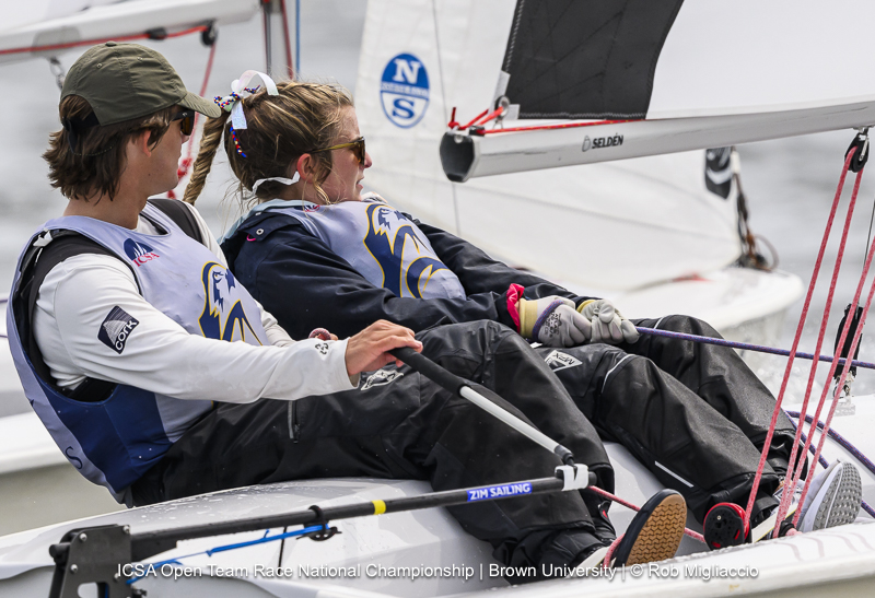 Two sailors in life vests and sunglasses steer a dinghy during a race. Both are focused on the water ahead, with ropes in hand, as the boat leans to one side. Race branding and sponsor logos are visible on their gear.