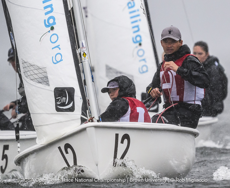 Two sailors in red and white vests steer a small sailboat marked with the number 12 during a regatta, with another boat close by. The water is choppy, and the sky appears overcast.