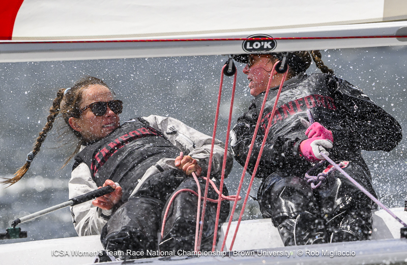 Two women in Stanford sailing gear compete in a race, leaning back on a boat as water sprays over them. Both are focused and wearing sunglasses, with ropes and sailing equipment visible.