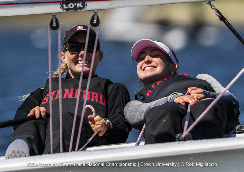 Two smiling women wearing Stanford gear sit in a sailboat during a race, leaning back and enjoying the moment, with blue water visible in the background. The image is from the ICSA Women’s Team Race National Championship.
