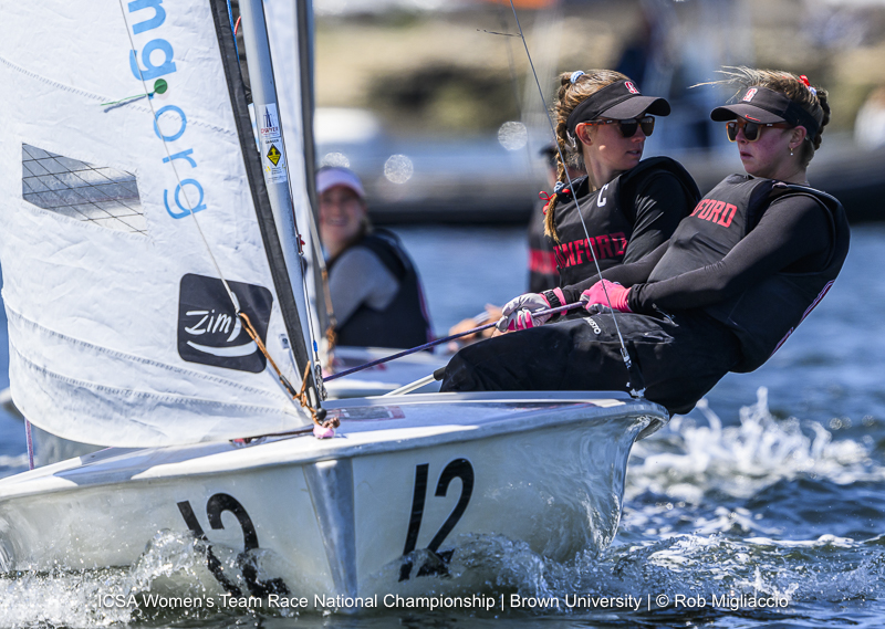 Two women sailors wearing sunglasses and Stanford gear are steering a small sailboat labeled '12' on the water during a race. Another sailor and boat are visible in the background.