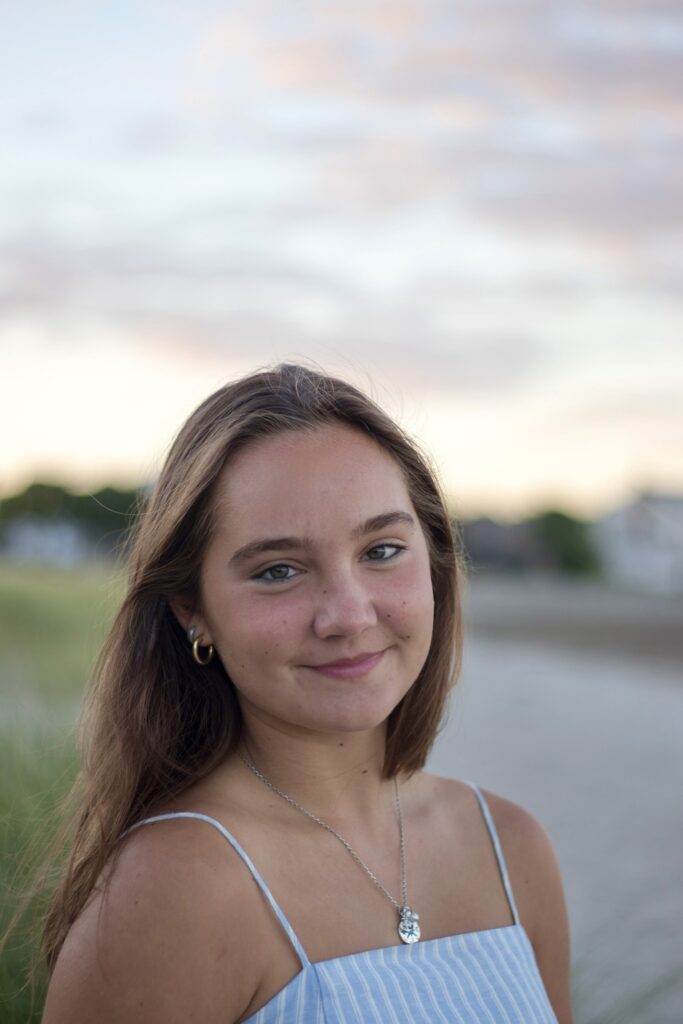 A young woman with long brown hair, wearing a blue and white striped spaghetti strap top and a silver necklace, smiles softly in an outdoor setting with blurred grass and sky in the background.