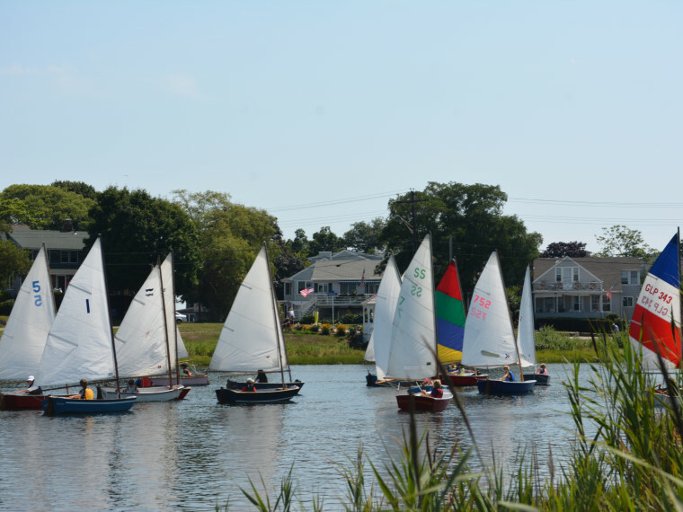 Several small sailboats with white sails are participating in a regatta on a calm river, with houses and greenery visible in the background under a clear blue sky.
