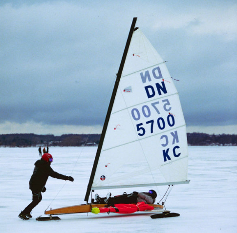 A person pushes an iceboat with a large sail and the markings 'DN 5700 KC' across a snowy, frozen lake, while another person lies inside the boat. Both are wearing helmets and winter clothing under a cloudy sky.