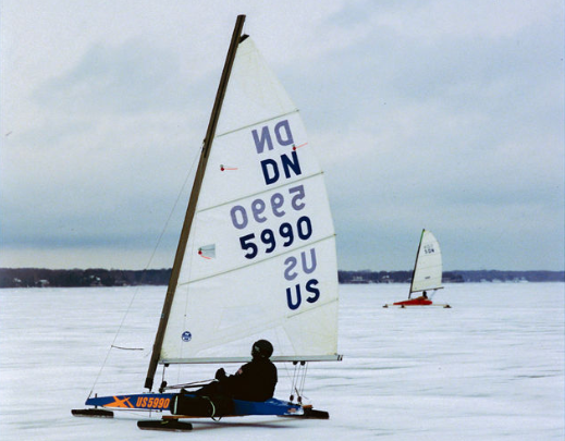 A person steers a small iceboat with a sail labeled "US 5990" across a frozen lake, while another iceboat sails in the distance under a cloudy sky.