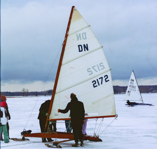 Several people stand around a DN-class iceboat on a frozen lake, while another iceboat sails in the distance under a cloudy sky. The ground is covered with snow and ice.