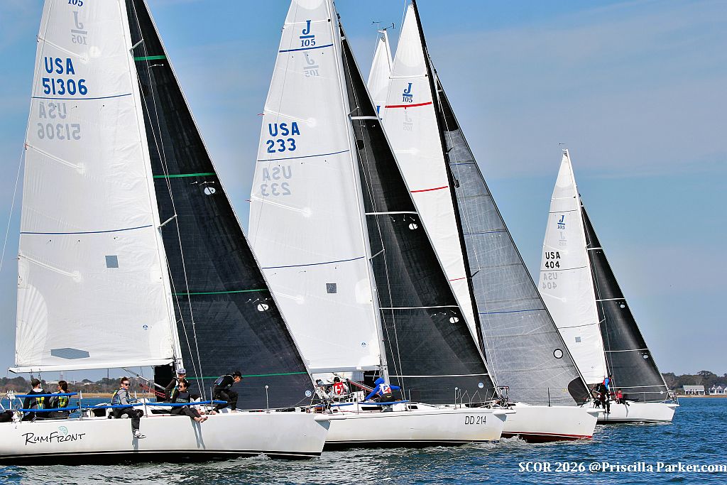 Several sailboats with white and black sails, each marked with "USA" and numbers, race closely together on calm water under a blue sky. People are visible on the boats, actively steering and managing sails.