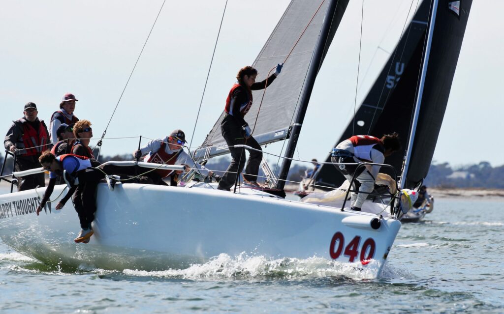 A sailboat with the number 040 and several crew members in life jackets racing on the water, leaning and working together as nearby boats compete in the background on a sunny day.
