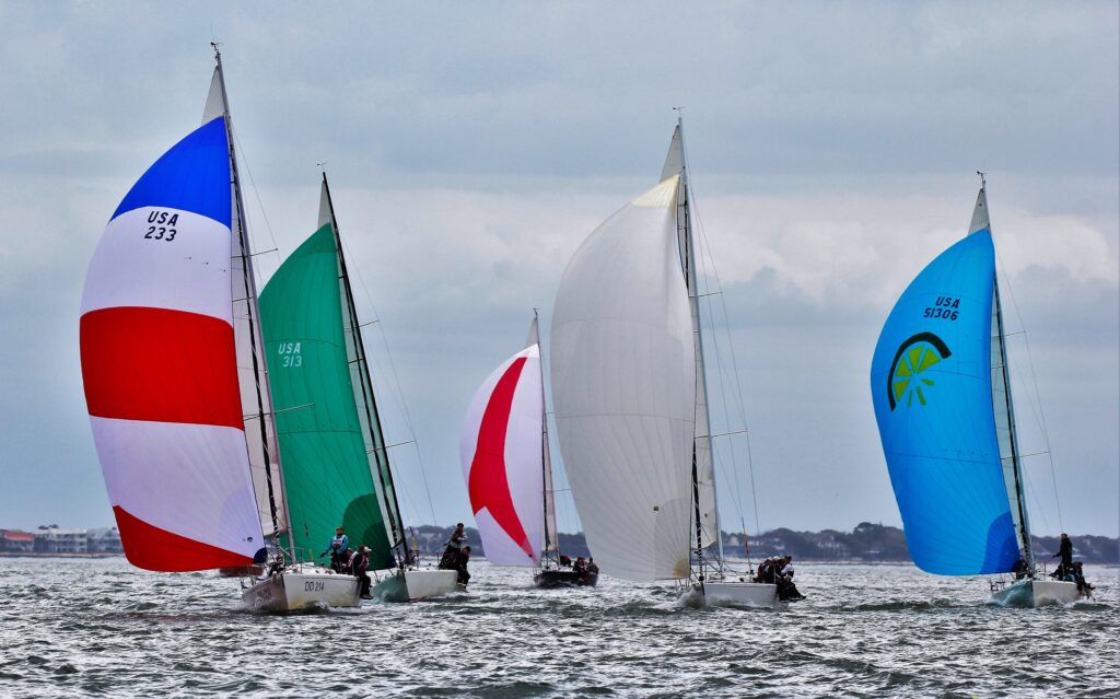 Four sailboats with colorful spinnaker sails—red, blue, green, and white—race on a choppy body of water under a cloudy sky, with a distant shoreline visible in the background.