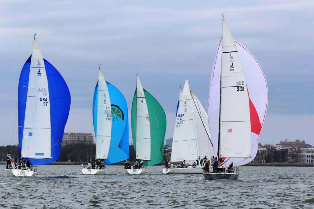 Six sailboats with colorful sails compete on the water near the shore, with buildings and trees visible in the background under a cloudy sky.