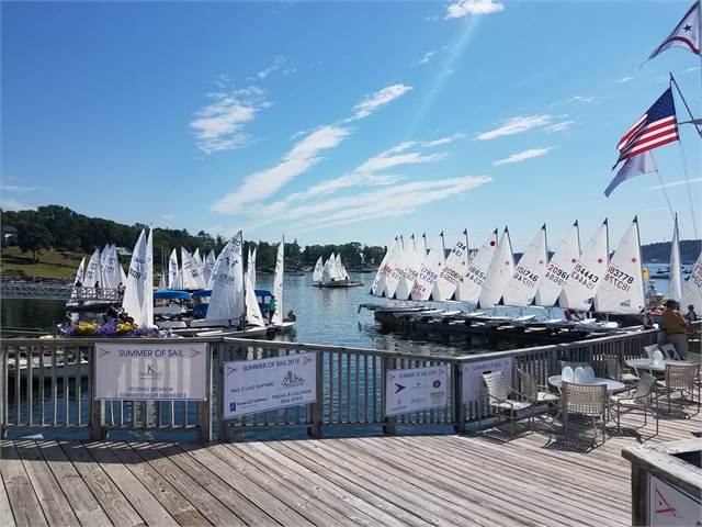 Dozens of small sailboats are docked by a wooden pier on a sunny day. Several white sails are visible, and American flags wave above. Outdoor chairs and tables sit on the deck in the foreground.