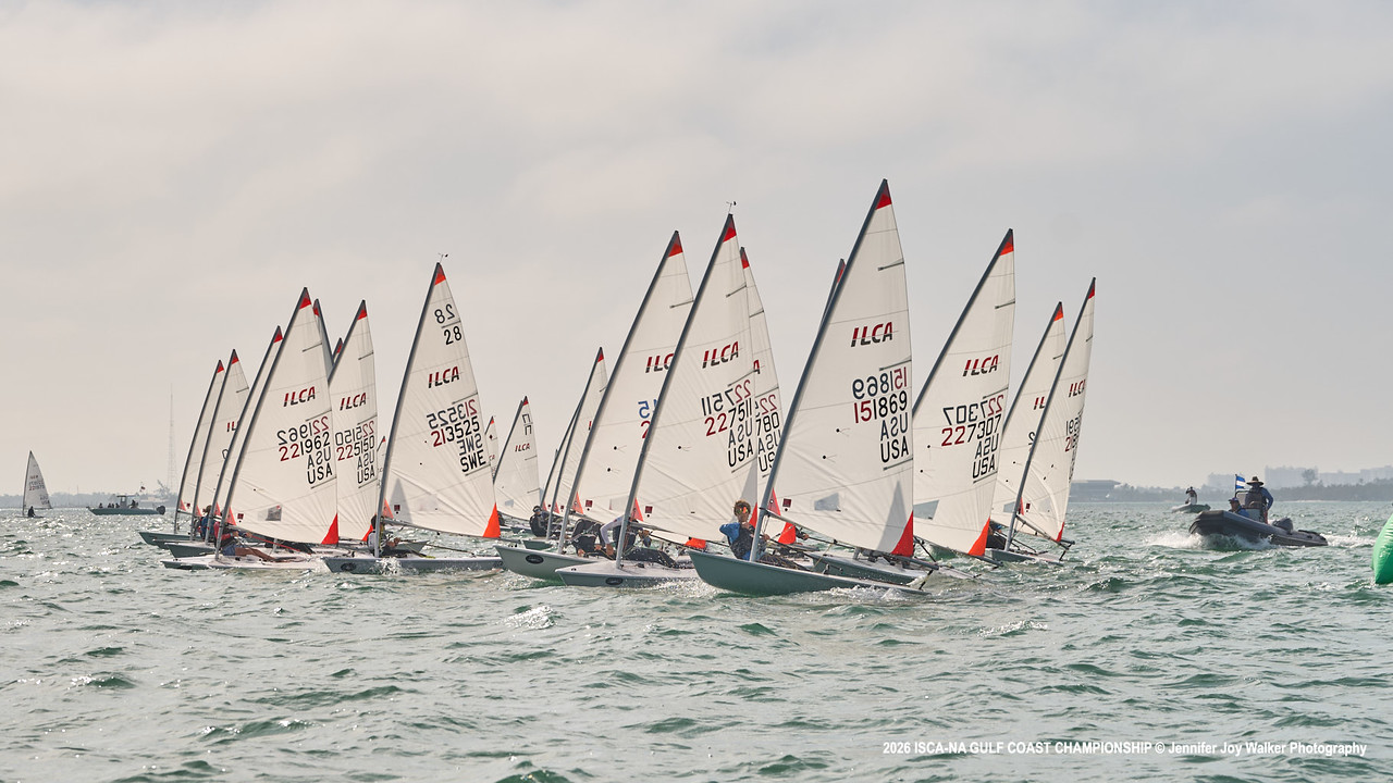 A fleet of small sailboats, each with white sails marked "ILCA," race closely together on a slightly choppy sea under a cloudy sky, accompanied by a motorboat on the right.