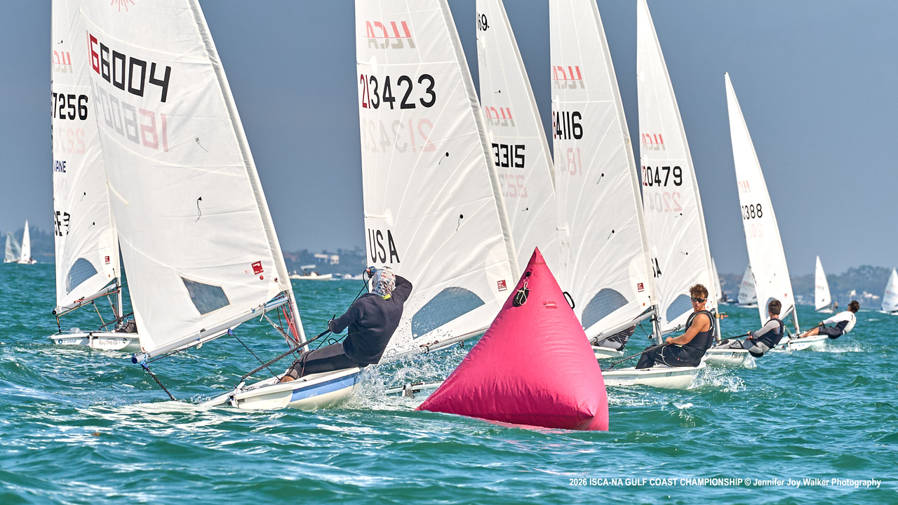 Several small sailboats race around a bright pink buoy on turquoise water. Sailors lean out to balance their boats under clear skies, with more boats visible in the background.