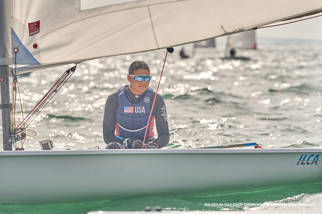 A sailor wearing a navy wetsuit with a USA flag and sunglasses sits in a sailboat on the water, holding the tiller. The sailboat is labeled "ILCA" and the background is sunlit with other boats visible.