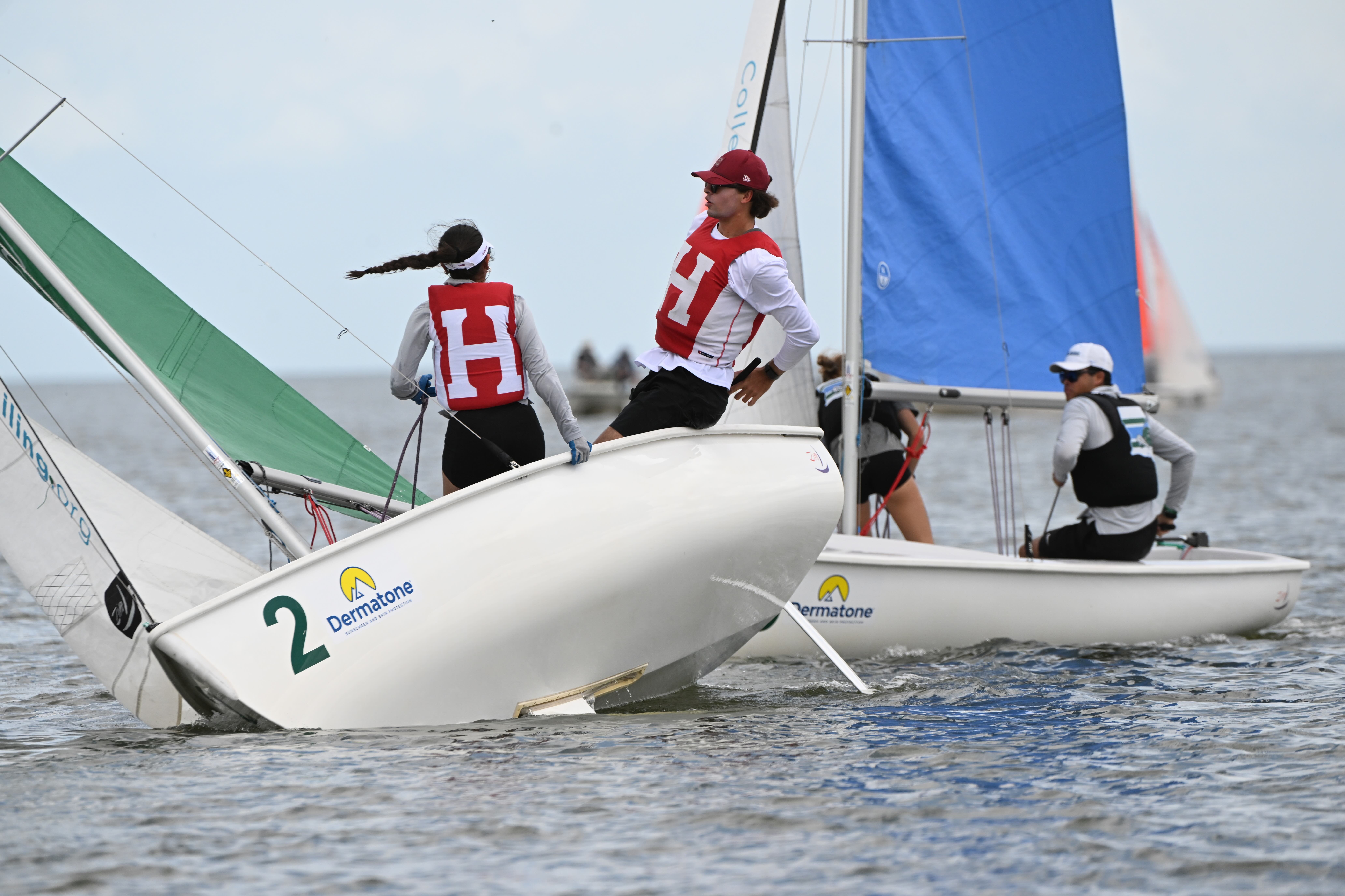Two sailboats race on the water; two people wearing red vests with the letter "H" are on one boat leaning to balance it, while two sailors in white and blue vests are on the other. The boats display "Dermatone" logos.