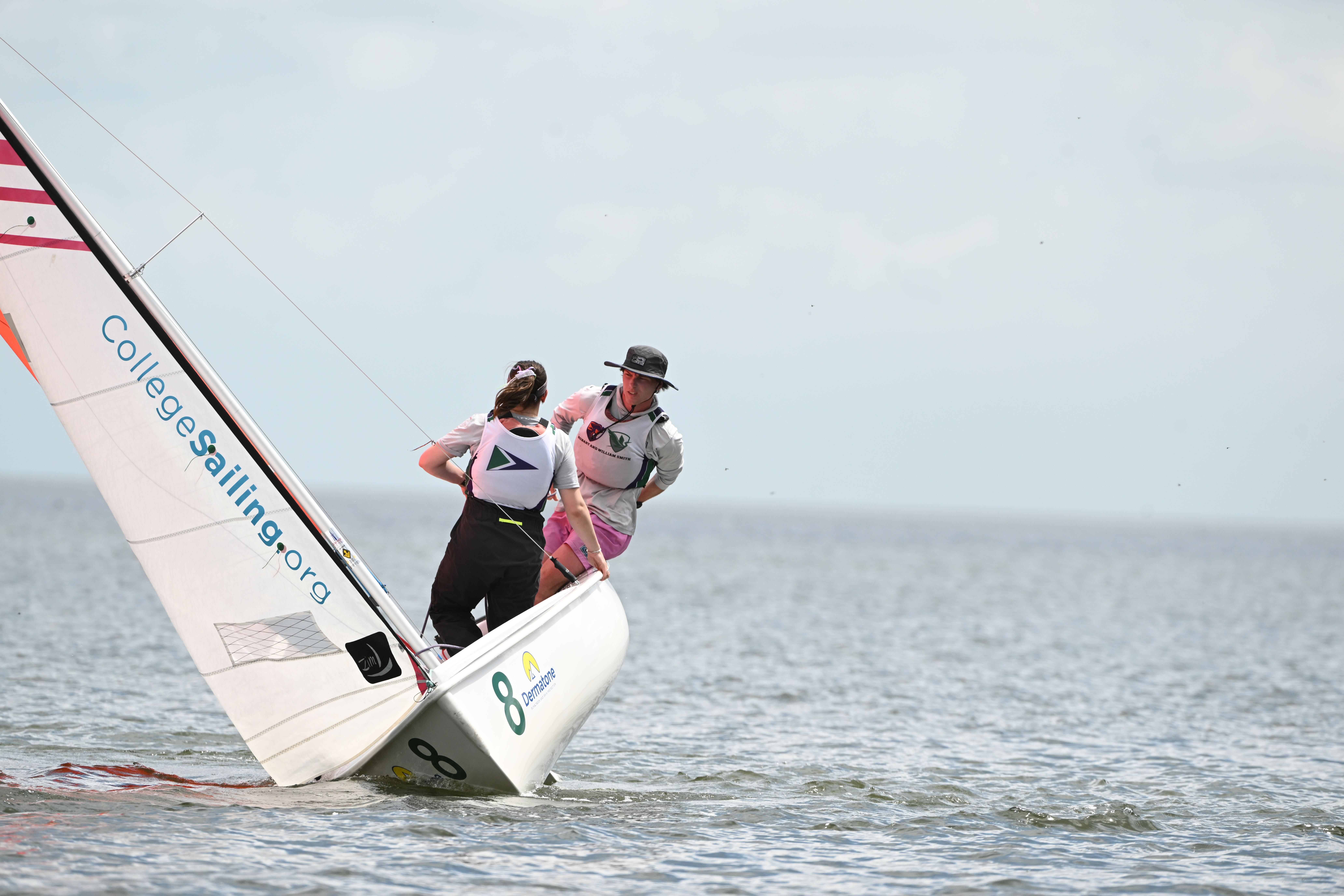 Two sailors in life jackets navigate a small sailboat with "CollegeSailing.org" on the sail. The boat tilts on the water, and the sailors lean out for balance under a bright, partly cloudy sky.