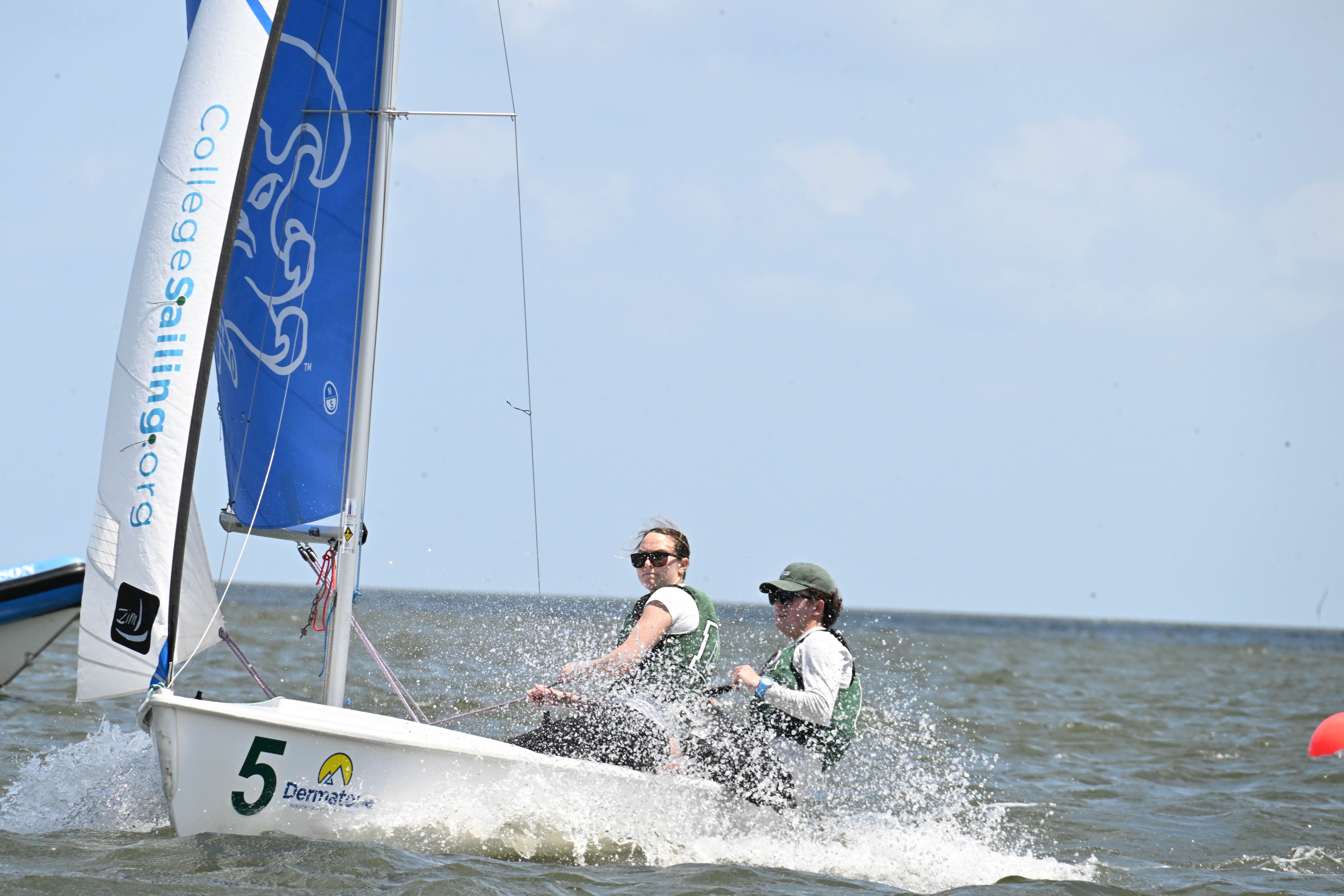 Two people wearing life jackets sail a small boat with a blue and white sail marked "CollegeSailing.org" through choppy water. The boat is numbered 5 and water splashes up at the front.